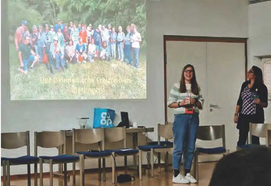 Zwei Frauen stehen vor einer Gruppe von Stühlen und präsentieren beim Herbstfest des Freizeitclubs, im Hintergrund eine Projektion mit einem Gruppenfoto.
